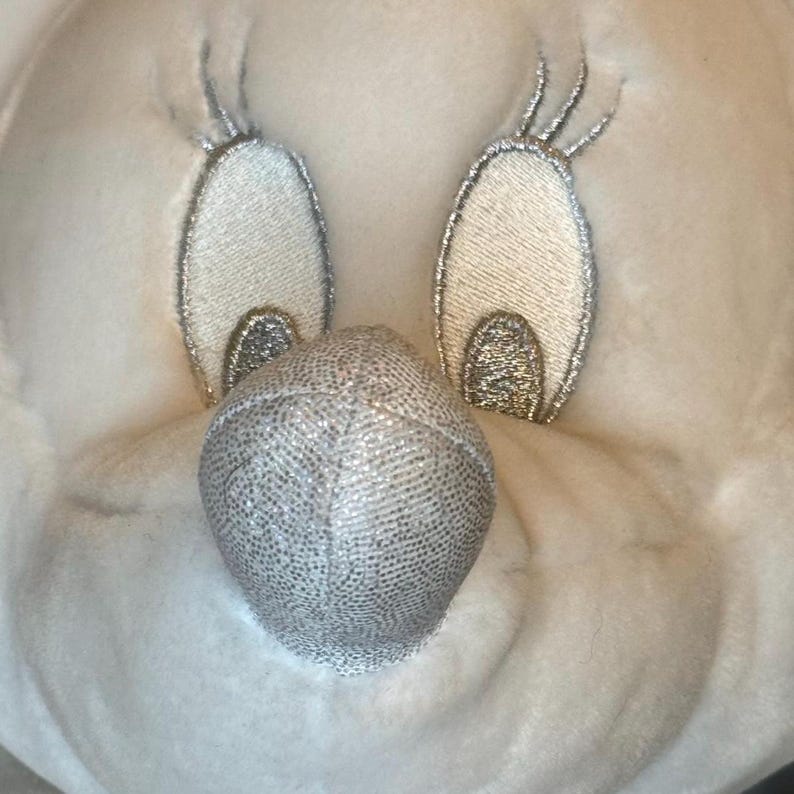 Puede incluir: Primer plano de una cara de peluche con detalles bordados. Los ojos son de forma ovalada con contornos y acentos de purpurina plateada. La nariz es una bola de purpurina plateada. El juguete est&aacute; hecho de un material suave y blanquecino.
