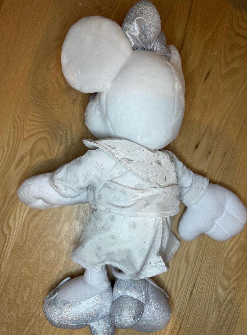 Puede incluir: Mu&ntilde;eca de peluche blanca de Minnie Mouse con lazo, vestido y zapatos plateados. El vestido tiene un estampado de copos de nieve. Se ven las orejas y las manos grandes de la mu&ntilde;eca. La mu&ntilde;eca mide unos 30 cm de alto.