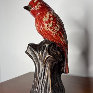 May include: A ceramic bird figurine in a vibrant red hue with gold accents, perched atop a dark brown tree stump. The bird has a detailed feather design and a black beak. This decorative piece adds a touch of nature-inspired art to any space.