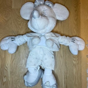Puede incluir: Un mu&ntilde;eco de peluche blanco de Minnie Mouse con detalles plateados. El mu&ntilde;eco viste un atuendo blanco con detalles de copos de nieve, una chaqueta a juego y un lazo plateado. Los zapatos del mu&ntilde;eco tambi&eacute;n tienen lazos plateados. El mu&ntilde;eco tiene orejas grandes y redondas y una cara sonriente.