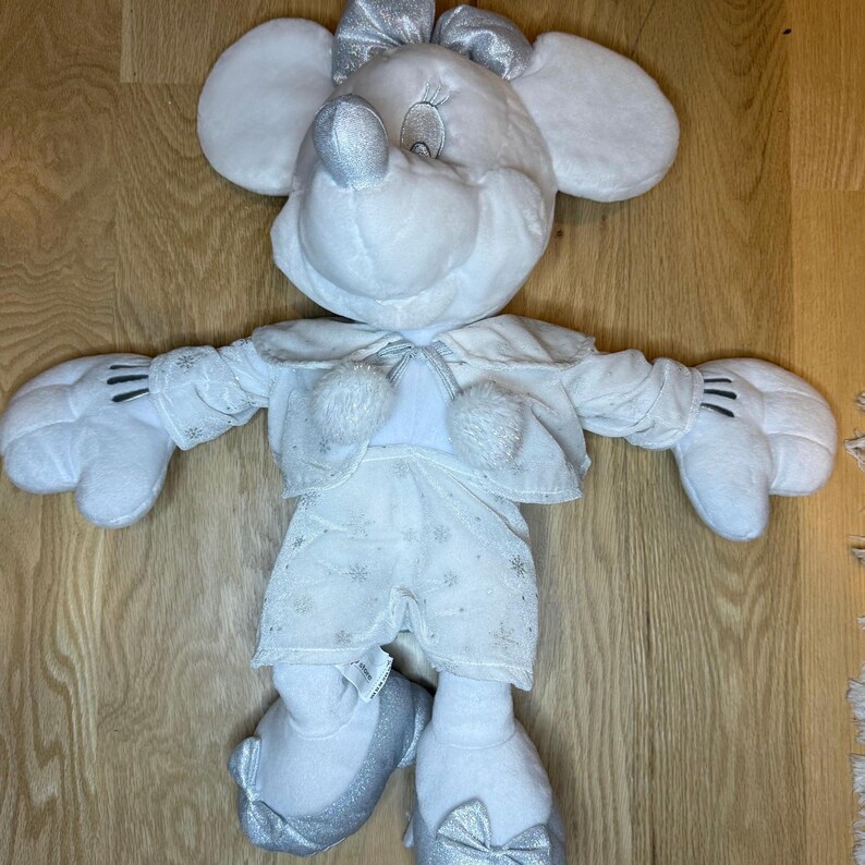 Puede incluir: Un mu&ntilde;eco de peluche blanco de Minnie Mouse con detalles plateados. El mu&ntilde;eco viste un atuendo blanco con detalles de copos de nieve, una chaqueta a juego y un lazo plateado. Los zapatos del mu&ntilde;eco tambi&eacute;n tienen lazos plateados. El mu&ntilde;eco tiene orejas grandes y redondas y una cara sonriente.