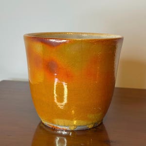 May include: A handcrafted ceramic pot with a glossy, textured finish. The pot features a gradient of colors, transitioning from a deep orange at the base to a reddish-brown at the rim. The interior is a lighter shade of orange.