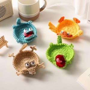 Peut inclure: Quatre bols en crochet circulaires présentant des motifs de créatures de style dessin animé en bleu, jaune, beige et vert. Chaque bol possède des traits distincts, incluant des yeux en plastique, des bouches ouvertes et des membres.