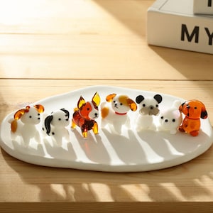May include: A collection of small, colorful glass dog figurines displayed on a white oval plate. The dogs feature various colors, including white, orange, black, and brown. The figurines are arranged on a wooden surface.