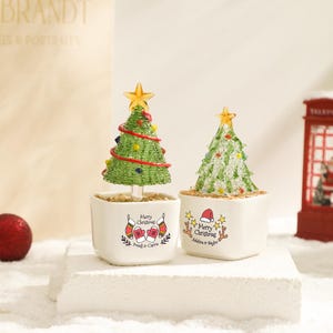 May include: Two miniature glass Christmas trees in square white ceramic pots. The left tree is green with red garland and colorful ornaments. The right tree is clear with green accents. Both pots feature "Merry Christmas" text and festive graphics.