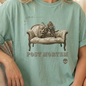 May include: A light green t-shirt with rolled-up sleeves featuring a graphic design. The design includes two carved vegetable faces on a miniature couch, the text "POST MORTEM", and a skull.