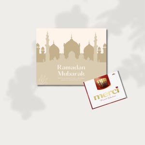 Puede incluir: Una tarjeta beige de felicitación de Ramadán Mubarak con una silueta de mezquita y el texto "Ramadán Mubarak". También hay una caja de bombones Merci con borde rojo y letras doradas.