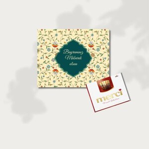 Puede incluir: Una tarjeta cuadrada con un patrón floral y el texto "Bayramınız Mübarek olsun" en un marco verde. Una pequeña caja de bombones "merci" está al lado de la tarjeta.