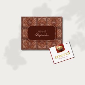Puede incluir: Una caja de regalo cuadrada con un diseño estampado y el texto "Hayırlı Bayramlar" en un rectángulo marrón. También se ve una caja más pequeña con la palabra "merci" en letras doradas y las palabras "Finest Selection".