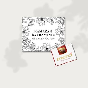 Puede incluir: Una tarjeta cuadrada blanca con ilustraciones florales negras y el texto "RAMAZAN BAYRAMINIZ MÜBAREK OLSUN". Una pequeña caja de bombones "merci" roja y blanca está al lado de la tarjeta.
