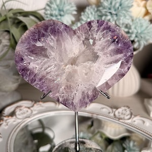 Könnte beinhalten: Eine herzförmige Amethyst-Geodenscheibe mit tiefvioletten und durchscheinenden weißen Kristallstrukturen. Der polierte Stein ist auf einem dünnen, silberfarbenen Metallständer mit rundem Fuß montiert und zeigt eine natürliche, raue Textur.