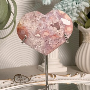 K&ouml;nnte beinhalten: Ein herzf&ouml;rmiger Amethyst-Geode auf einem silbernen Metallst&auml;nder. Der Geode zeigt eine Mischung aus lila, rosa und wei&szlig;en Kristallen. Der St&auml;nder hat einen runden Sockel und eine gebogene Halterung. Dekorative Blumen sind im Hintergrund.