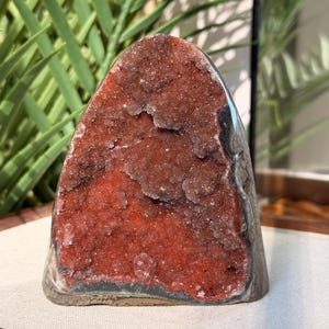 May include: A geode crystal with a reddish-brown interior and a gray and white exterior. The crystal has a rounded top and a flat base. The interior is filled with small, sparkling crystals.