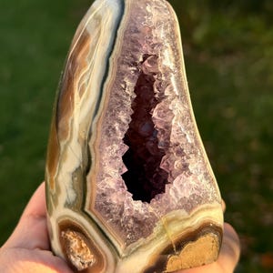 May include: A polished amethyst geode, displaying a cavity filled with sparkling purple crystals. The exterior has bands of brown, white, and green. The geode is held in a hand, highlighting its natural beauty.