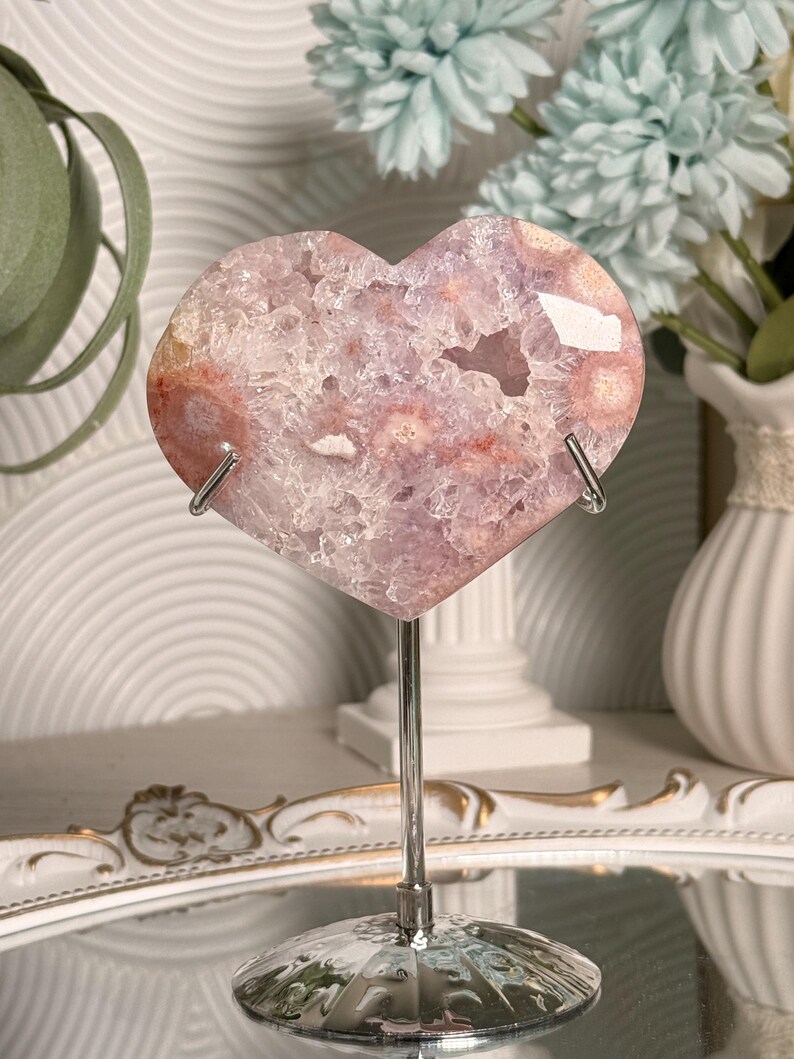 K&ouml;nnte beinhalten: Ein herzf&ouml;rmiger Amethyst-Geode auf einem silbernen Metallst&auml;nder. Der Geode zeigt eine Mischung aus lila, rosa und wei&szlig;en Kristallen. Der St&auml;nder hat einen runden Sockel und eine gebogene Halterung. Dekorative Blumen sind im Hintergrund.