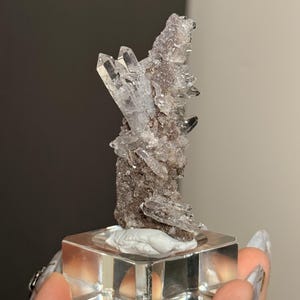 Pu&ograve; includere: Un gruppo di cristalli di quarzo trasparenti con terminazioni appuntite e affilate, fissato su una matrice minerale scura e ruvida. L'esemplare &egrave; montato in verticale su una base quadrata in acrilico trasparente, mostrando formazioni naturali.