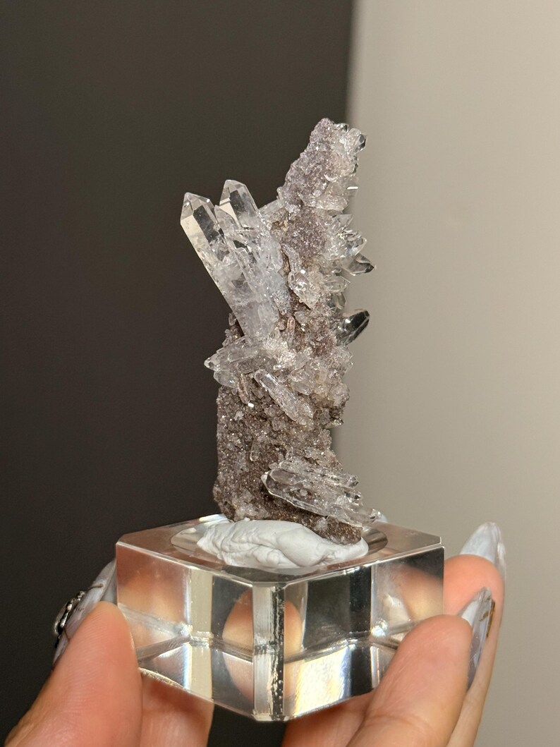 Pu&ograve; includere: Un gruppo di cristalli di quarzo trasparenti con terminazioni appuntite e affilate, fissato su una matrice minerale scura e ruvida. L'esemplare &egrave; montato in verticale su una base quadrata in acrilico trasparente, mostrando formazioni naturali.