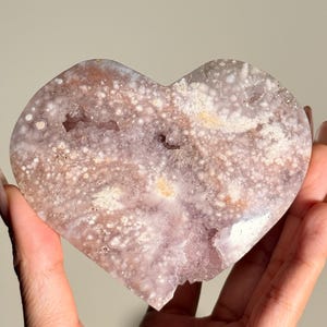 Puede incluir: Geoda de amatista en forma de coraz&oacute;n con una superficie rugosa y texturizada. El cristal presenta una mezcla de tonos lavanda, blancos y marrones, con peque&ntilde;as formaciones cristalinas. La geoda se sostiene en una mano.
