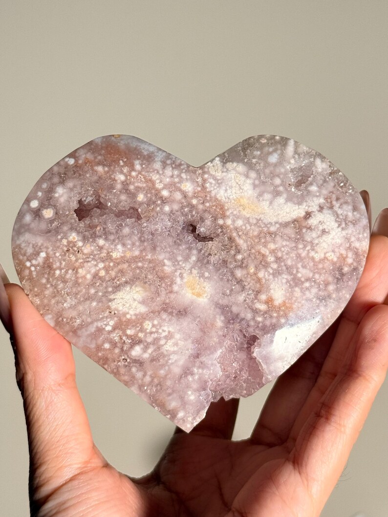 Puede incluir: Geoda de amatista en forma de coraz&oacute;n con una superficie rugosa y texturizada. El cristal presenta una mezcla de tonos lavanda, blancos y marrones, con peque&ntilde;as formaciones cristalinas. La geoda se sostiene en una mano.