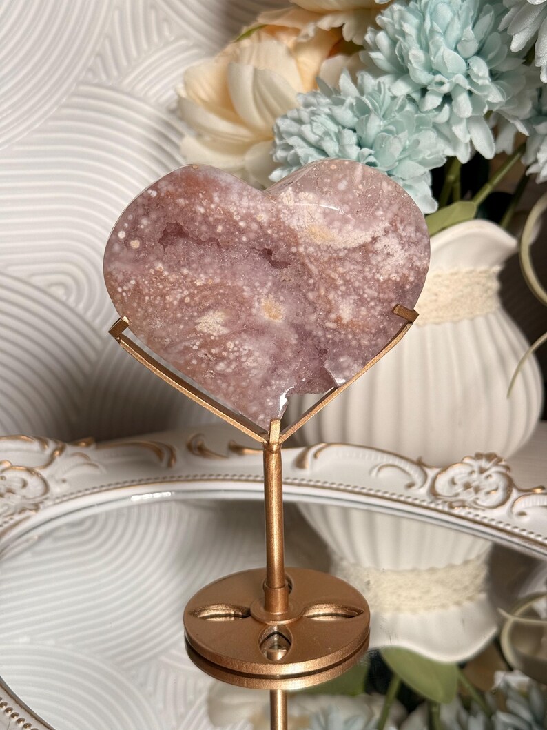 Puede incluir: Una geoda de amatista en forma de coraz&oacute;n sobre un soporte de metal dorado. La geoda tiene una mezcla de colores morados y blancos con una textura cristalina. El soporte tiene una base circular y un dise&ntilde;o decorativo. Art&iacute;culo decorativo.