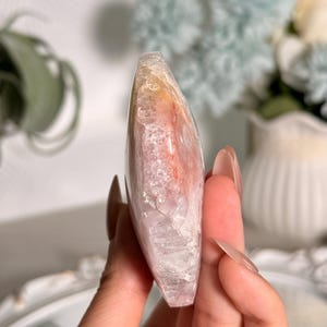K&ouml;nnte beinhalten: Ein polierter, tropfenf&ouml;rmiger Kristall mit einer Mischung aus Farben. Der Stein hat ein durchscheinendes Aussehen mit Farbt&ouml;nen von Rosa, Orange und Wei&szlig;. Der Kristall wird in einer Hand gehalten und zeigt seine glatte, reflektierende Oberfl&auml;che.
