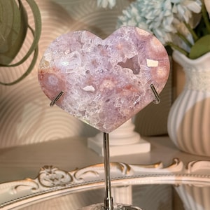 K&ouml;nnte beinhalten: Ein herzf&ouml;rmiger, polierter Kristallgeode in Rosa-, Lila- und Wei&szlig;t&ouml;nen, montiert auf einem silbernen Metallst&auml;nder. Der Geode hat ein kristallines Inneres und wird vor einem verschwommenen Hintergrund mit Blumen pr&auml;sentiert.