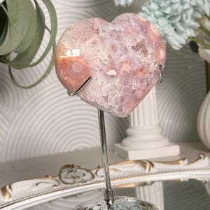 K&ouml;nnte beinhalten: Ein herzf&ouml;rmiger Kristallgeode in Rosa- und Wei&szlig;t&ouml;nen, montiert auf einem silbernen Metallst&auml;nder. Der Geode hat eine strukturierte Oberfl&auml;che und wird vor einem verschwommenen Hintergrund mit dekorativen Elementen pr&auml;sentiert.