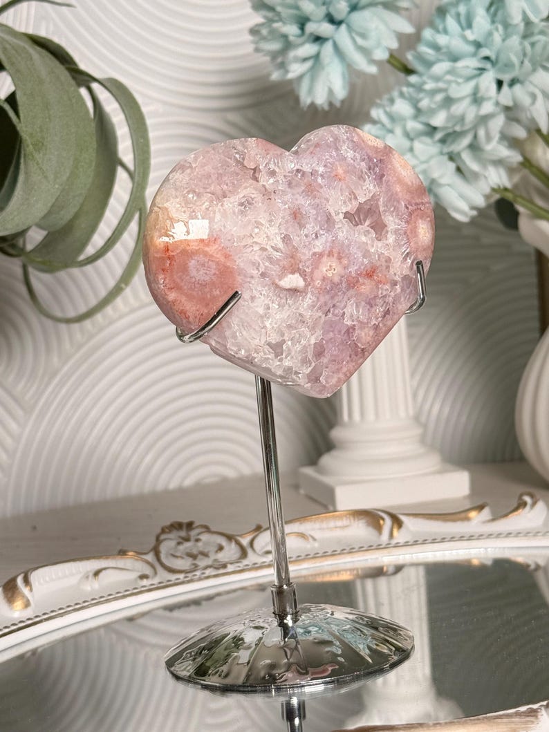 K&ouml;nnte beinhalten: Ein herzf&ouml;rmiger Kristallgeode in Rosa- und Wei&szlig;t&ouml;nen, montiert auf einem silbernen Metallst&auml;nder. Der Geode hat eine strukturierte Oberfl&auml;che und wird vor einem verschwommenen Hintergrund mit dekorativen Elementen pr&auml;sentiert.