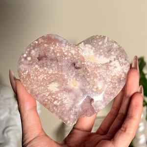 Puede incluir: Un cristal en forma de coraz&oacute;n con un patr&oacute;n moteado en tonos rosa, blanco y marr&oacute;n. El cristal se sostiene en una mano, mostrando su textura natural y variaciones de color. Art&iacute;culo decorativo.