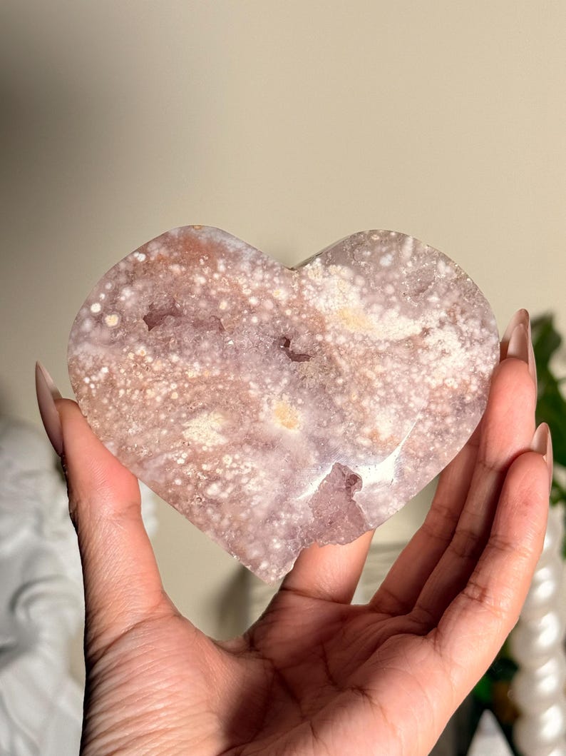 Puede incluir: Un cristal en forma de coraz&oacute;n con un patr&oacute;n moteado en tonos rosa, blanco y marr&oacute;n. El cristal se sostiene en una mano, mostrando su textura natural y variaciones de color. Art&iacute;culo decorativo.