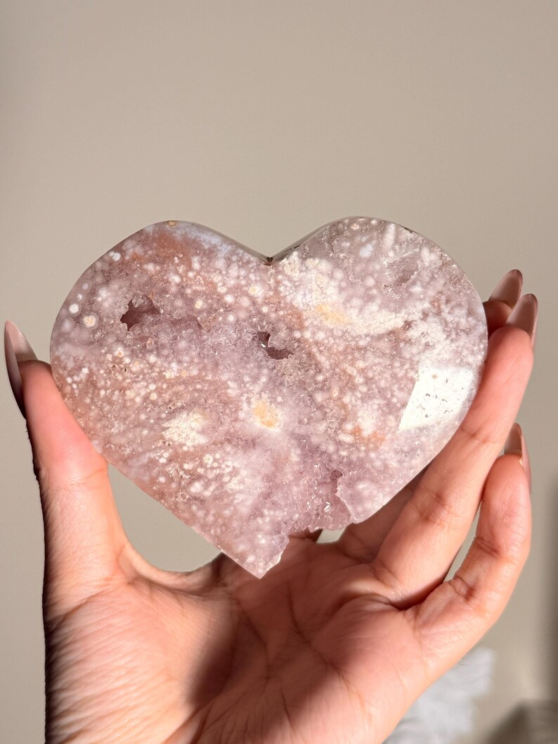 Puede incluir: Geoda de cristal en forma de coraz&oacute;n en tonos rosa, blanco y marr&oacute;n. La geoda tiene una superficie rugosa y texturizada con estructuras cristalinas visibles y un centro hueco. El cristal se sostiene en una mano.
