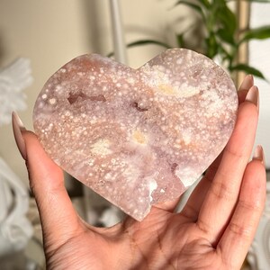 Puede incluir: Geoda de cristal en forma de coraz&oacute;n con una superficie moteada, de color p&uacute;rpura claro y blanco. La geoda tiene un aspecto rugoso y texturizado y se sostiene en una mano. El cristal tiene una forma natural y org&aacute;nica y es un objeto decorativo.