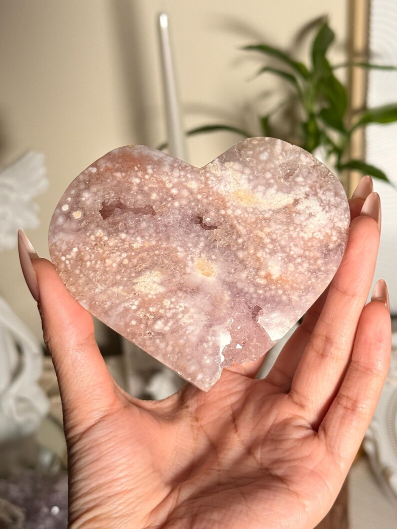 Puede incluir: Geoda de cristal en forma de coraz&oacute;n con una superficie moteada, de color p&uacute;rpura claro y blanco. La geoda tiene un aspecto rugoso y texturizado y se sostiene en una mano. El cristal tiene una forma natural y org&aacute;nica y es un objeto decorativo.