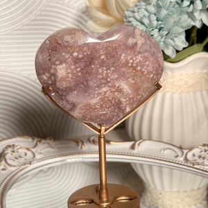 Puede incluir: Una piedra pulida en forma de coraz&oacute;n, en tonos lavanda y blanco, montada sobre un soporte de metal dorado. La piedra tiene un patr&oacute;n moteado y se exhibe frente a un jarr&oacute;n blanco con flores.