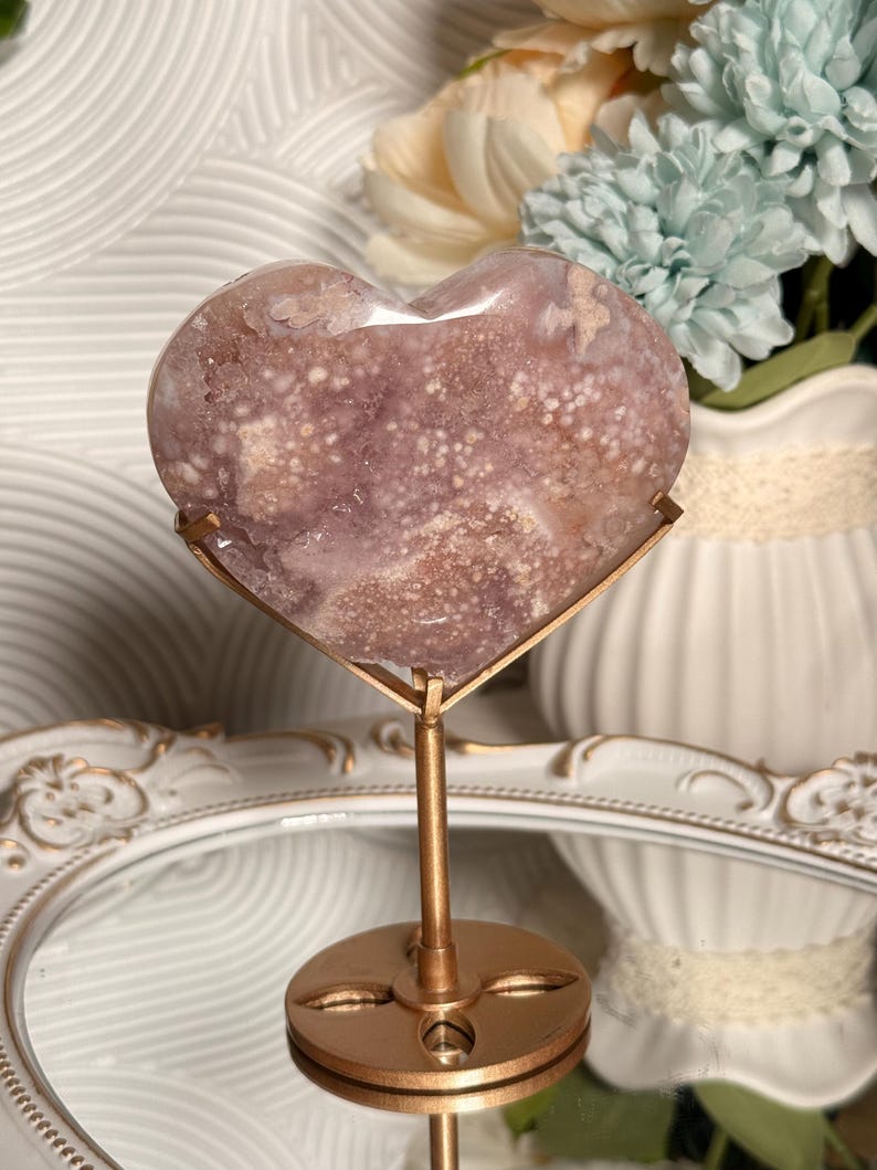 Puede incluir: Una piedra pulida en forma de coraz&oacute;n, en tonos lavanda y blanco, montada sobre un soporte de metal dorado. La piedra tiene un patr&oacute;n moteado y se exhibe frente a un jarr&oacute;n blanco con flores.