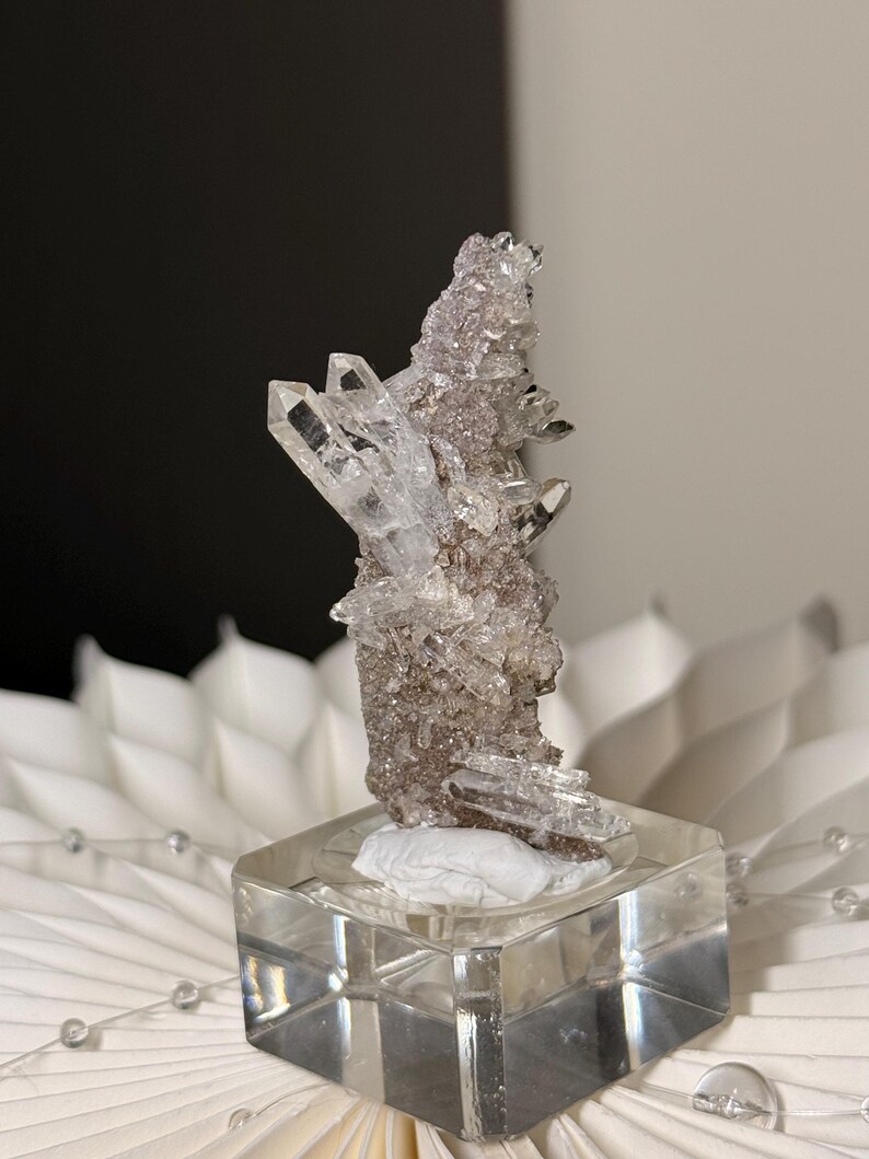 Pu&ograve; includere: Un grappolo di cristalli di quarzo trasparenti e appuntiti con una base a matrice testurizzata color marrone-grigio. Il campione minerale &egrave; montato su un supporto quadrato in acrilico trasparente, che mette in mostra molteplici punte cristalline.