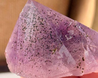 Può includere: Primo piano di un cristallo viola traslucido sfaccettato con inclusioni verde scuro. Il cristallo ha una forma triangolare con una punta. Lo sfondo è sfocato e di tono neutro.