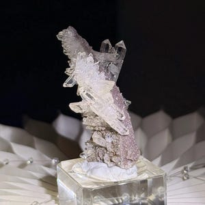 Pu&ograve; includere: Un grappolo di cristalli di quarzo trasparenti e appuntiti che crescono su una base minerale testurizzata color rosa polvere. L'esemplare &egrave; montato su un supporto quadrato in vetro trasparente, che mette in risalto la struttura cristallina.