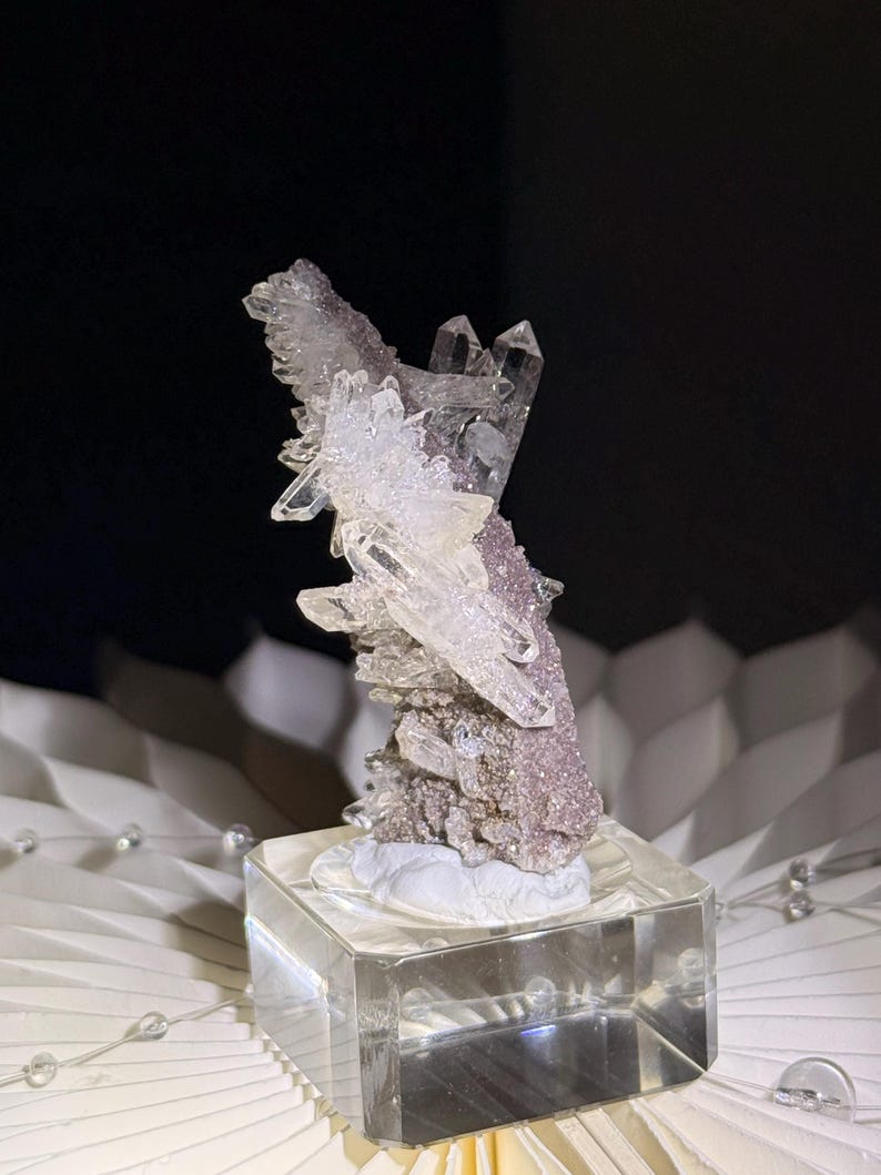 Pu&ograve; includere: Un grappolo di cristalli di quarzo trasparenti e appuntiti che crescono su una base minerale testurizzata color rosa polvere. L'esemplare &egrave; montato su un supporto quadrato in vetro trasparente, che mette in risalto la struttura cristallina.