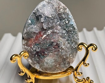 Op de afbeelding: Een geode kristallen ei met grijze, blauwe en rode tinten, tentoongesteld op een goudkleurige decoratieve standaard. De standaard heeft een sierlijk, opgerold ontwerp. Het eivormige kristal is het middelpunt.