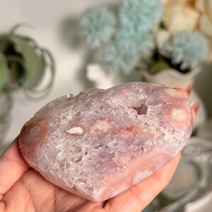 K&ouml;nnte beinhalten: Ein herzf&ouml;rmiger Kristall-Geode in Rosa- und Lavendelt&ouml;nen. Die Geode hat ein kristallines Inneres und eine glatte, polierte Au&szlig;enseite. Die Geode wird in einer Hand gehalten, wodurch ihre Gr&ouml;&szlig;e und Form hervorgehoben werden. Der Kristall hat ein nat&uuml;rliches, organisches Aussehen.
