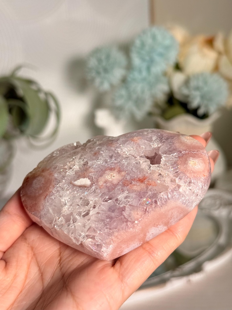 K&ouml;nnte beinhalten: Ein herzf&ouml;rmiger Kristall-Geode in Rosa- und Lavendelt&ouml;nen. Die Geode hat ein kristallines Inneres und eine glatte, polierte Au&szlig;enseite. Die Geode wird in einer Hand gehalten, wodurch ihre Gr&ouml;&szlig;e und Form hervorgehoben werden. Der Kristall hat ein nat&uuml;rliches, organisches Aussehen.
