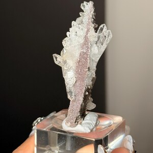 Pu&ograve; includere: Un esemplare minerale verticale caratterizzato da cristalli di quarzo trasparenti e appuntiti che crescono da una matrice centrale testurizzata di colore malva. L'esemplare &egrave; montato su una base quadrata in acrilico trasparente.