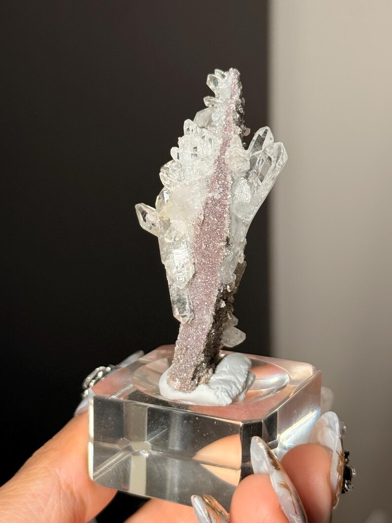 Pu&ograve; includere: Un esemplare minerale verticale caratterizzato da cristalli di quarzo trasparenti e appuntiti che crescono da una matrice centrale testurizzata di colore malva. L'esemplare &egrave; montato su una base quadrata in acrilico trasparente.