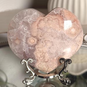 Puede incluir: Una piedra de ágata flor en forma de corazón, de tonos rosa y blanco, con estructuras cristalinas y patrones circulares visibles, expuesta sobre un soporte de metal plateado con patas curvas ornamentadas y base circular.