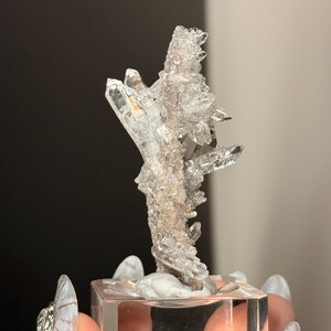Pu&ograve; includere: Un grappolo di cristalli di quarzo trasparenti e frastagliati con molteplici terminazioni appuntite e una superficie cristallina testurizzata. Il campione minerale &egrave; montato verticalmente su una base quadrata in acrilico trasparente, mostrando la sua struttura naturale, grezza e traslucida.