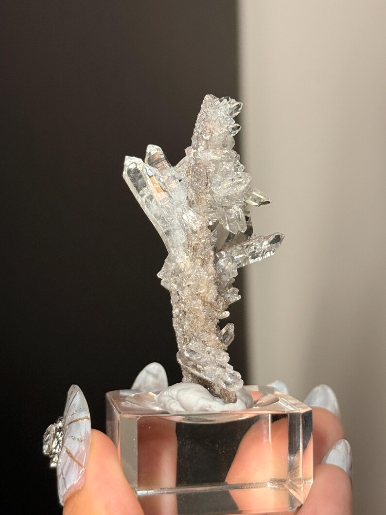 Pu&ograve; includere: Un grappolo di cristalli di quarzo trasparenti e frastagliati con molteplici terminazioni appuntite e una superficie cristallina testurizzata. Il campione minerale &egrave; montato verticalmente su una base quadrata in acrilico trasparente, mostrando la sua struttura naturale, grezza e traslucida.