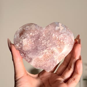 Puede incluir: Un cristal en forma de coraz&oacute;n, en tonos rosa y blanco, se sostiene en una mano. El cristal tiene una superficie texturizada con un patr&oacute;n moteado. La est&eacute;tica general es natural y org&aacute;nica, lo que sugiere un enfoque en cristales y piedras preciosas.