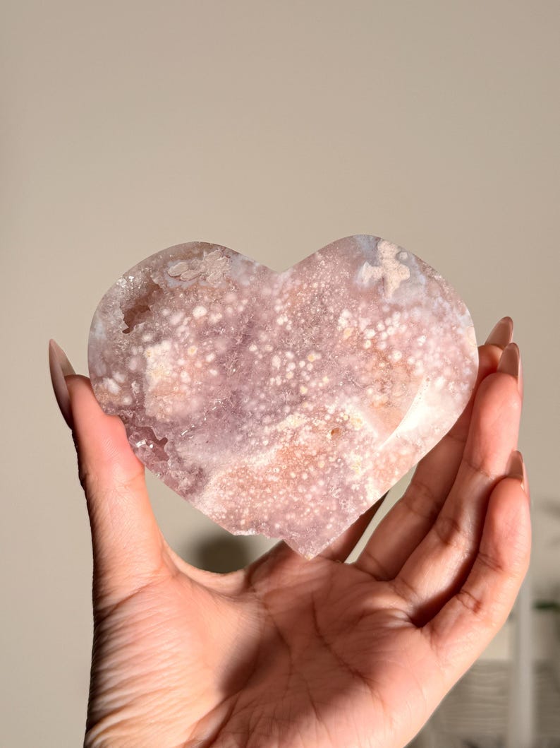 Puede incluir: Un cristal en forma de coraz&oacute;n, en tonos rosa y blanco, se sostiene en una mano. El cristal tiene una superficie texturizada con un patr&oacute;n moteado. La est&eacute;tica general es natural y org&aacute;nica, lo que sugiere un enfoque en cristales y piedras preciosas.