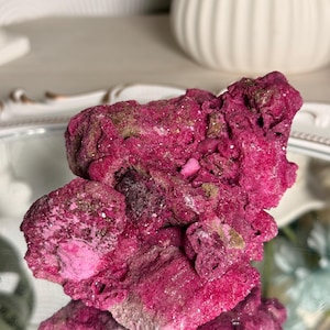 May include: A vibrant magenta mineral specimen with a rough, porous, and crystalline texture. The rock features deep pink hues with subtle metallic gold-toned flecks scattered across its uneven, jagged surface, reflecting light.